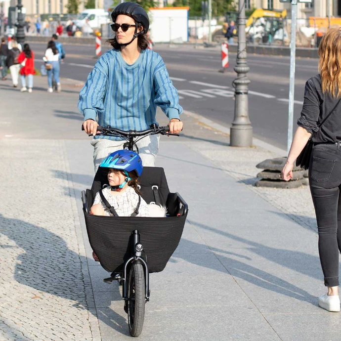 Eine Frau fährt ein muli Lastenrad auf einer Nebenanlage, vorne drin sitzt ein Kind.