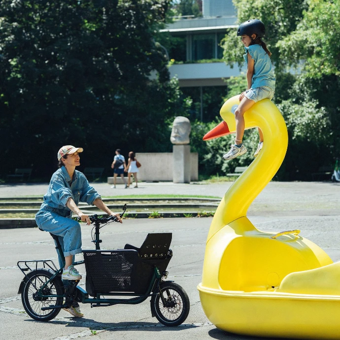 Eine Frau steht mit ihrem muli cycles vor einem gelben stillgelegten Tretboot in Enten/Schwanform, wo ein Kind auf dem Kopf sitzt